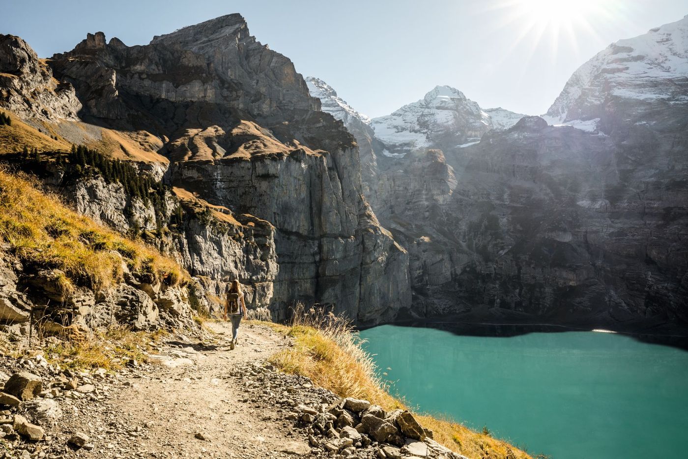 A hiker walks along a rugged, sunlit cliffside trail overlooking the vibrant turquoise waters of Lake Oeschinen near Kandersteg in the Swiss Alps