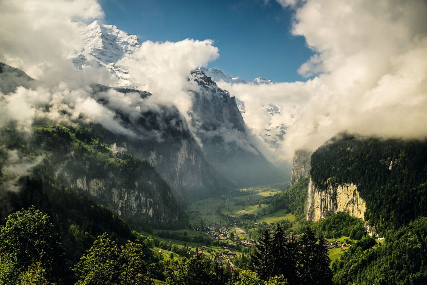 Clouds and mist weave through the towering, snow-dusted cliffs and lush green hillsides framing the spectacular Lauterbrunnen Valley in the Swiss Alps
