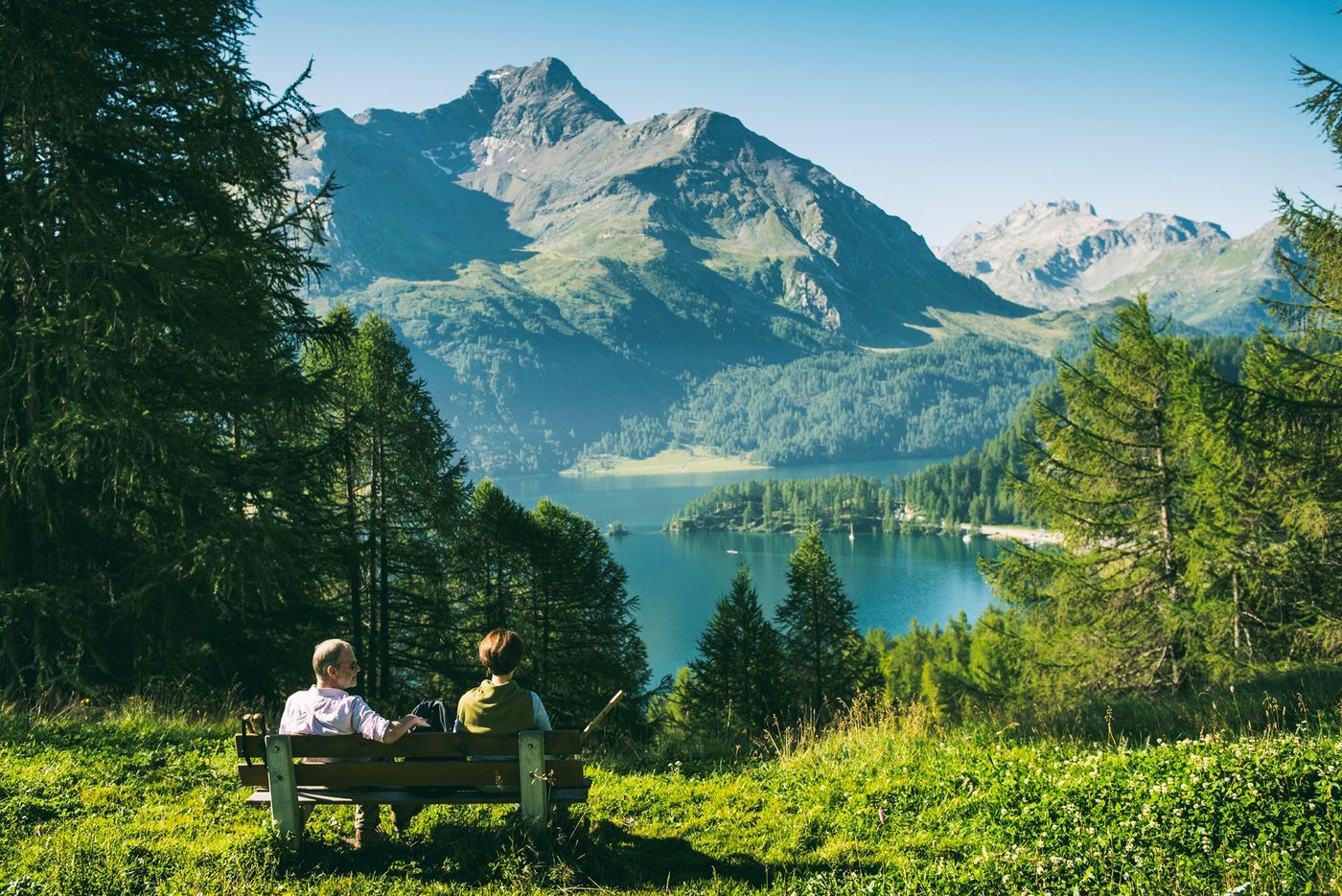 Two hikers resting on a wooden bench looking out over the clear blue waters of Lake Sils